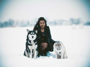 A woman kneeling in the snow flanked by two wolves on either side of her in the snow.